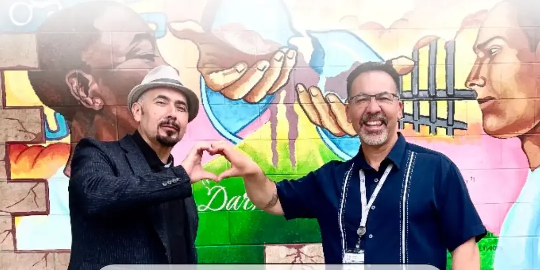 Two men forming a heart with their hands in front of a community mural